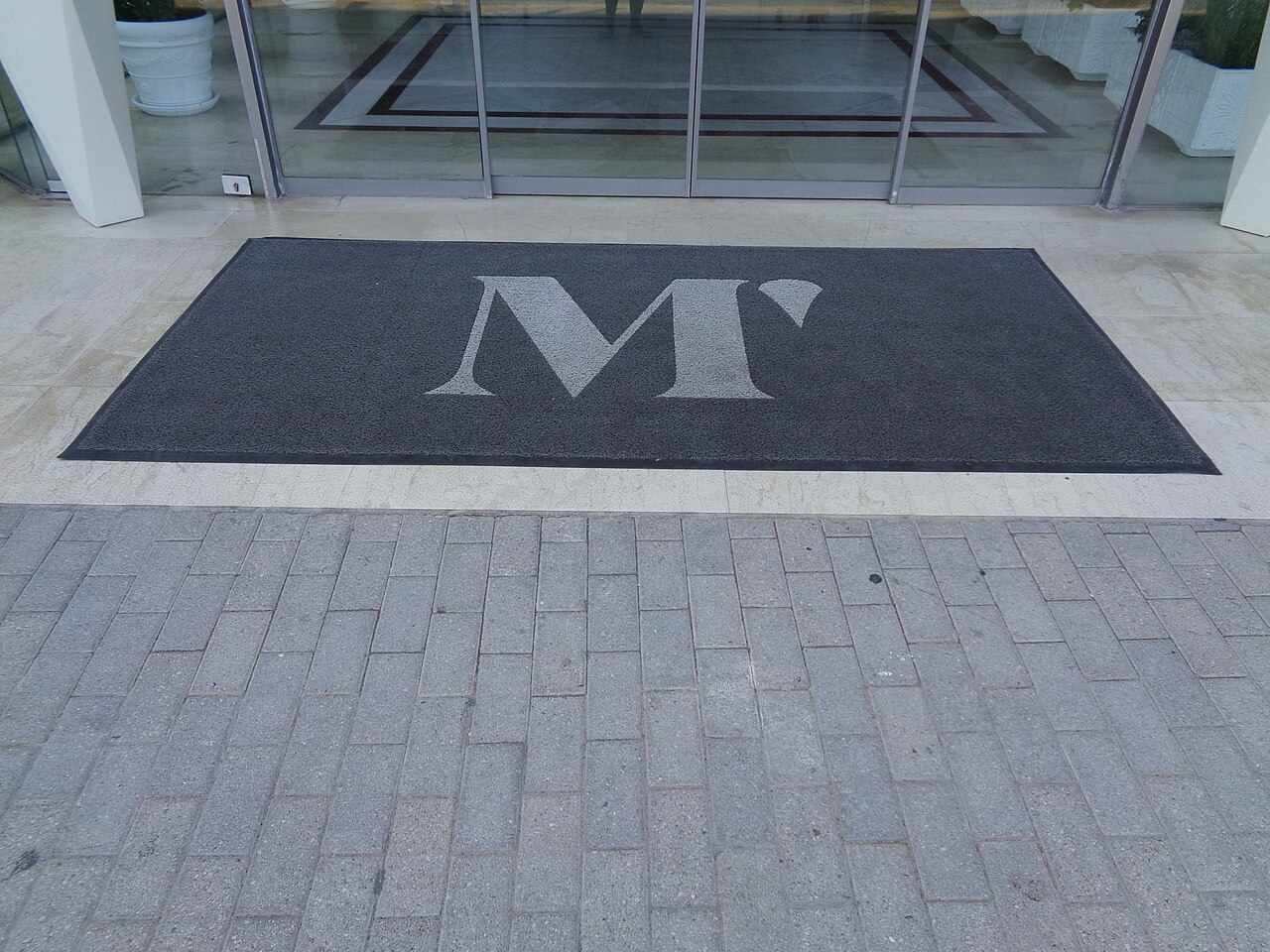 A dark entrance mat with a large white M mark before glass doors at a hotel.