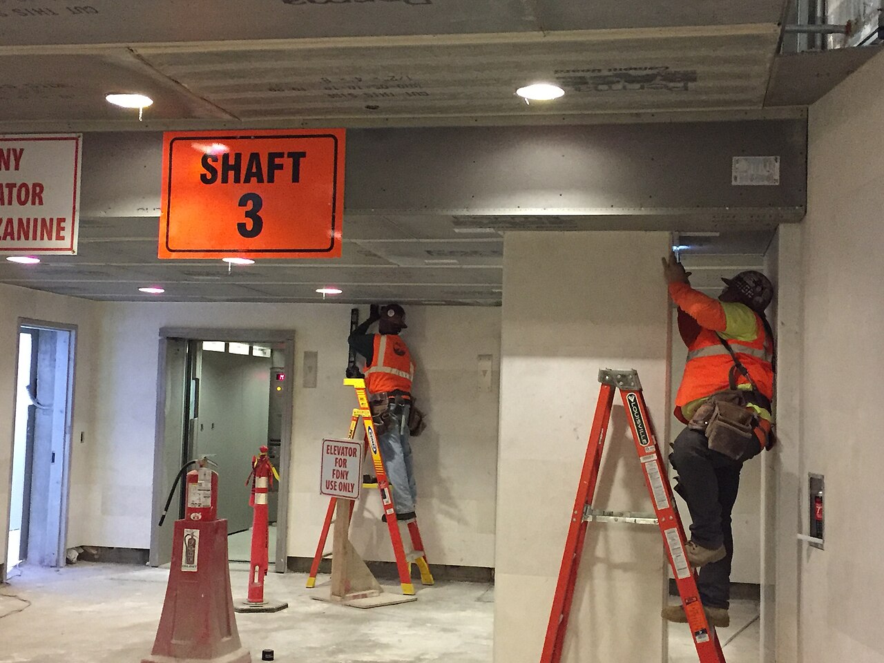 Carpenters installing cement board on a ceiling near an elevator lobby in a station concourse under construction