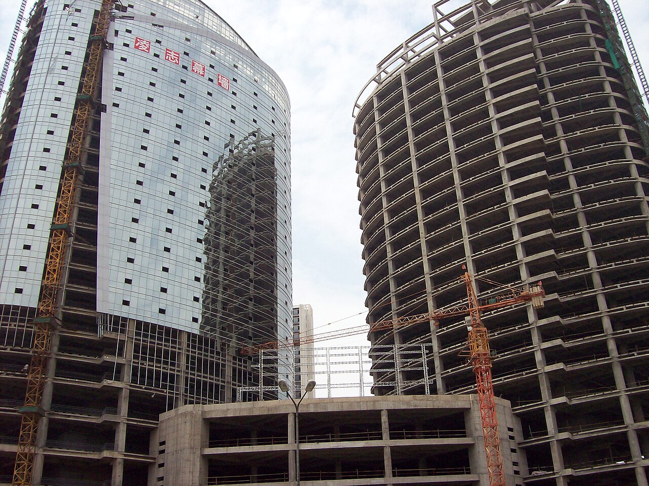Two towers under construction, one showing the structural frame and the other with its glass curtain wall installed