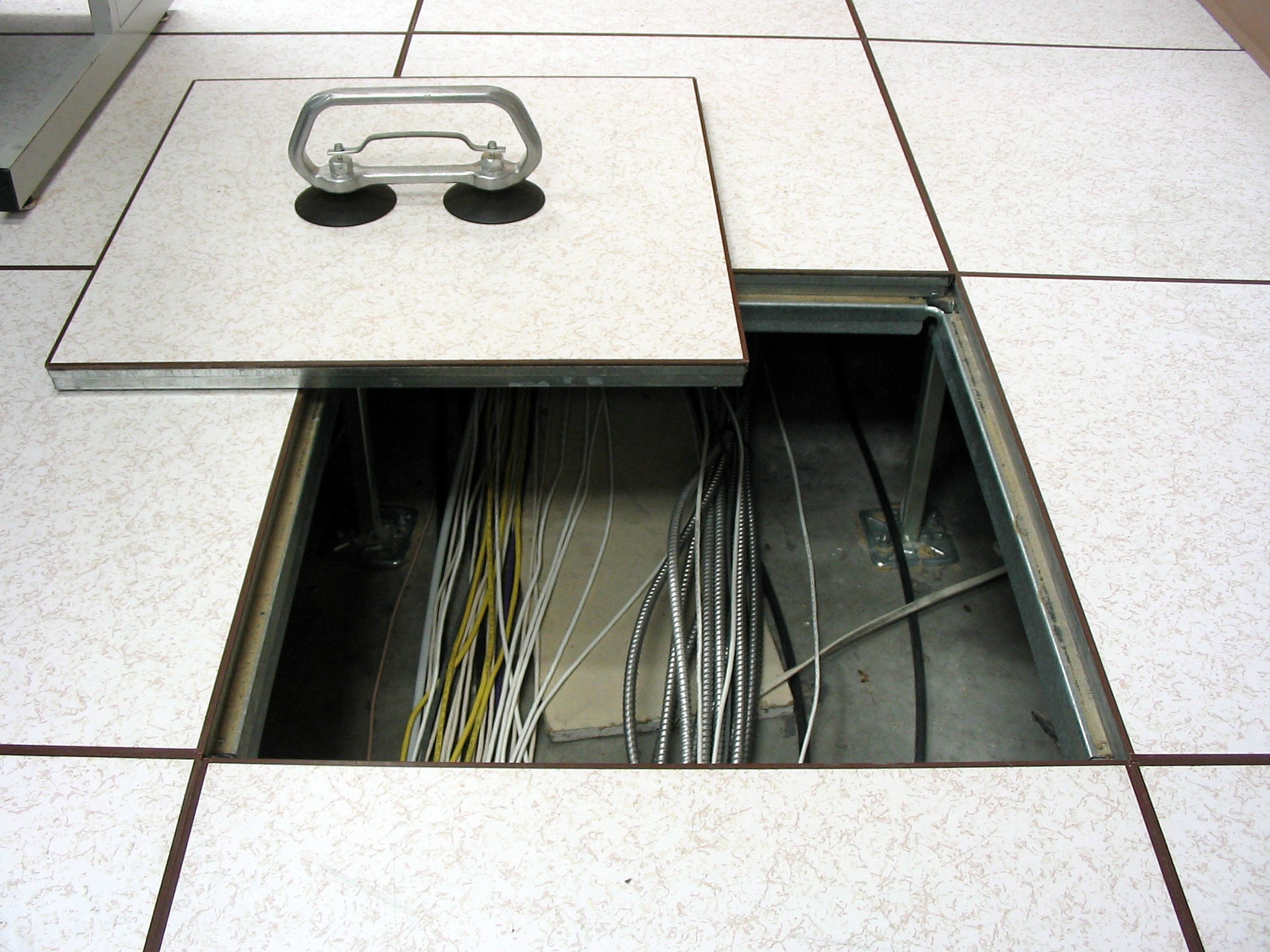 A raised floor panel lifted out with a suction lifter, showing cables and conduit in the void below