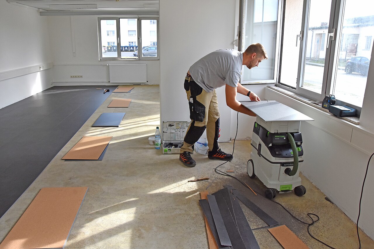 A worker installing vinyl flooring in a barracks day room during a renovation