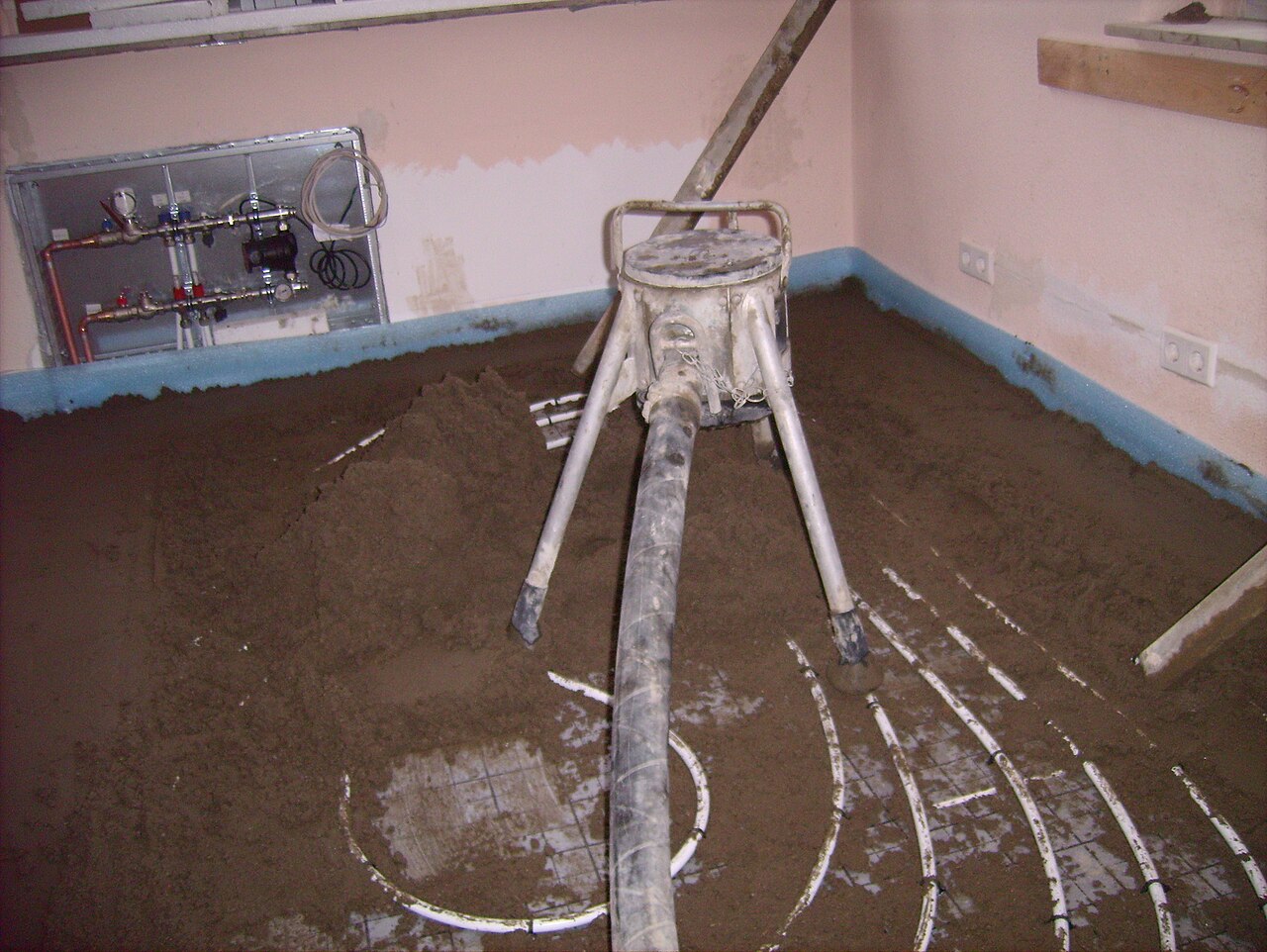 Screed being placed through a hose over underfloor heating pipes laid on insulation, with an edge insulation strip along the wall